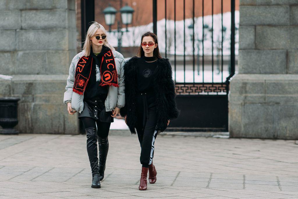 Two stylish women walk side by side in an urban setting, wearing black outfits with bold red accessories and sunglasses.