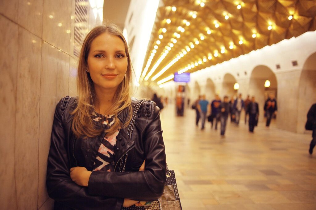 Woman with long blonde hair leaning against a marble wall in a busy, warmly lit station,