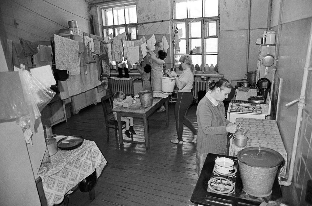 Busy communal kitchen with clothes hanging to dry, people washing dishes and cooking at a stove and table in the background.