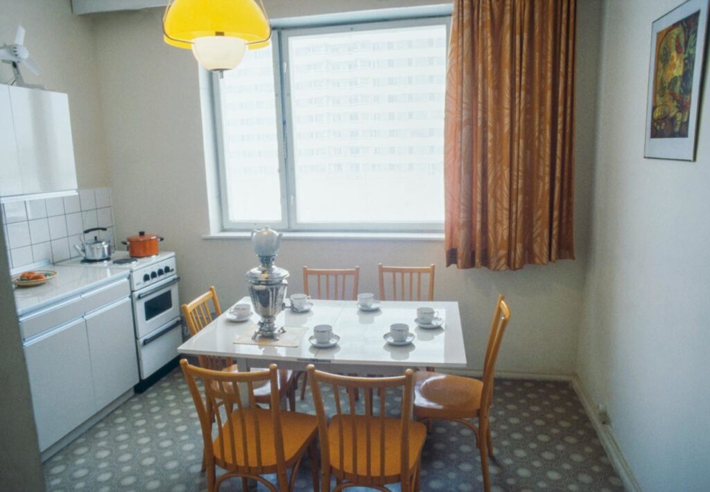 Dining nook with a white table set for six, a silver samovar centerpiece, and wooden yellow chairs under a yellow ceiling light by a large window with orange patterned curtains.