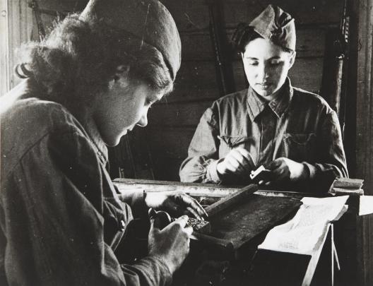 Two women in military uniforms work at a workbench, assembling or inspecting equipment together.