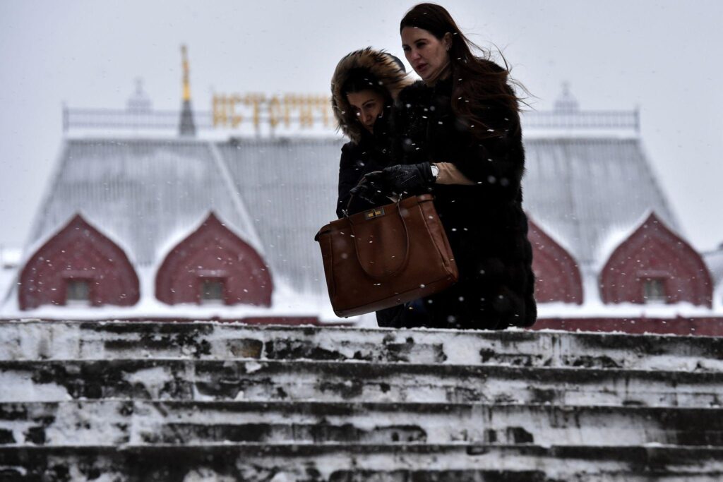 Two women in winter coats descend snow-covered steps, one carrying a large brown handbag.