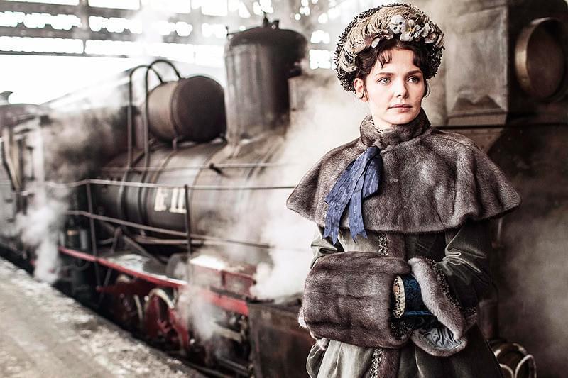 Victorian-era woman in a fur-trimmed coat and bonnet stands beside a steaming locomotive in a rail yard.